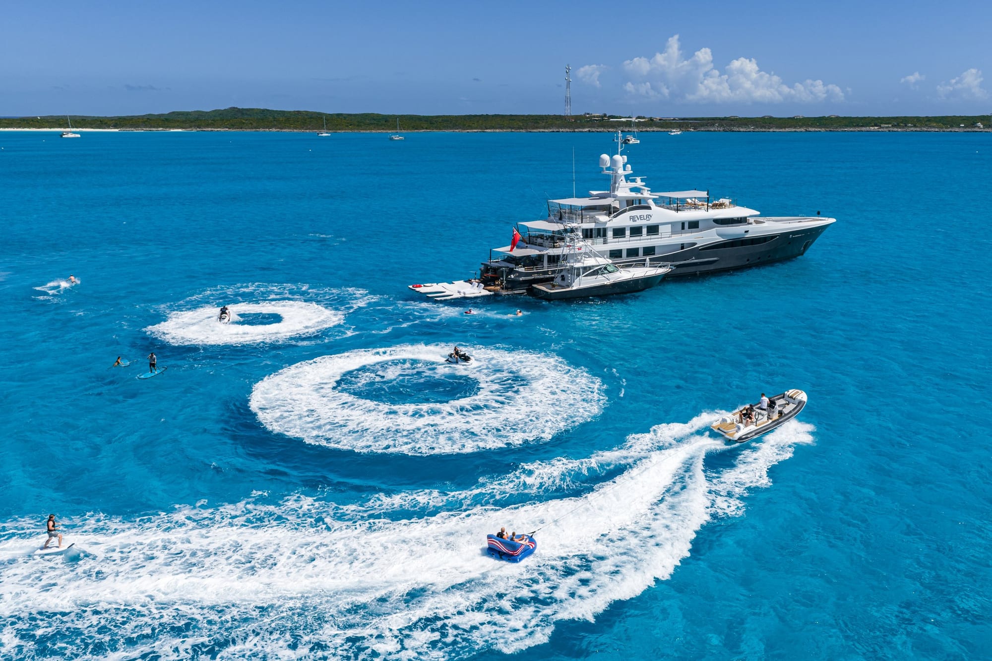 Aerial view of a superyacht at anchor in turquoise Bahamas water, with tenders and water toys circling