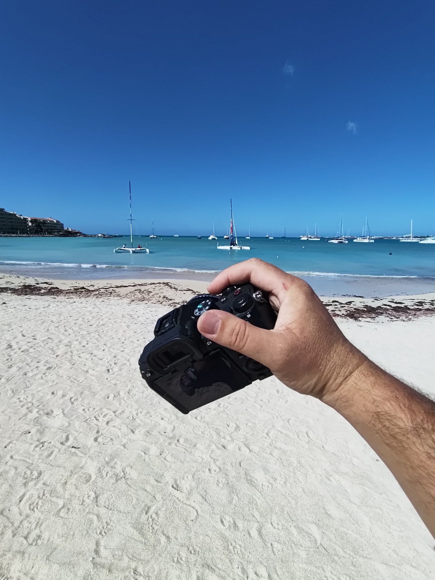 Camera held up over a white sand beach with yachts at anchor