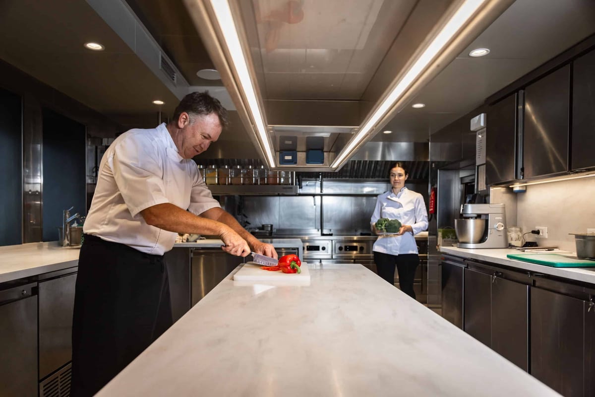 Two chefs preparing vegetables in a bright stainless steel yacht galley.