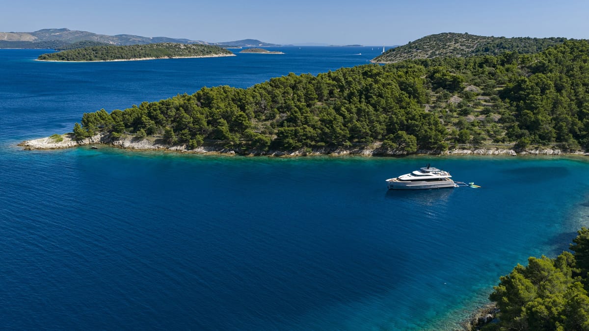 Motor yacht at anchor in deep blue water beside a pine-covered headland