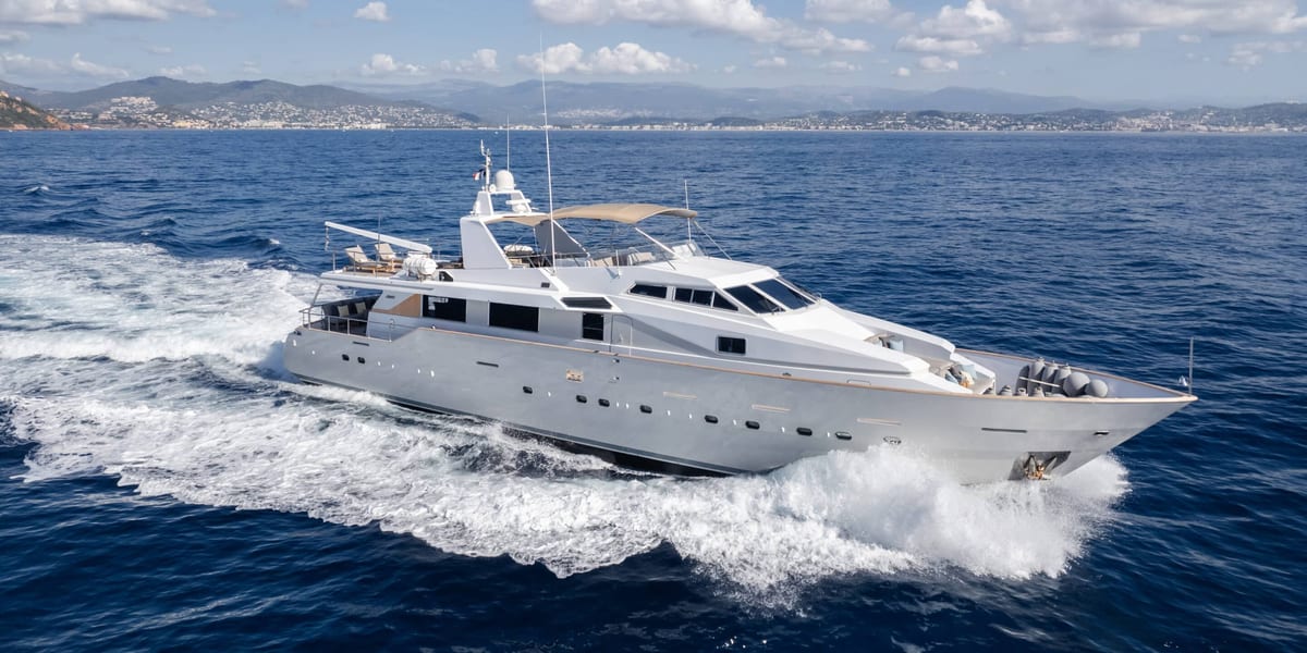 White motor yacht cruising at speed with a hilly coastline in the background.