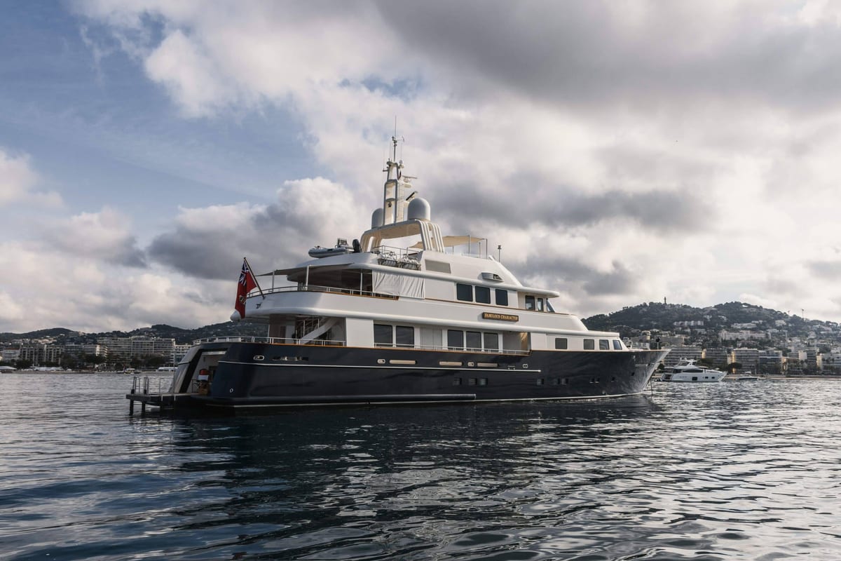 Superyacht with a dark hull and red ensign moored off a hillside town.