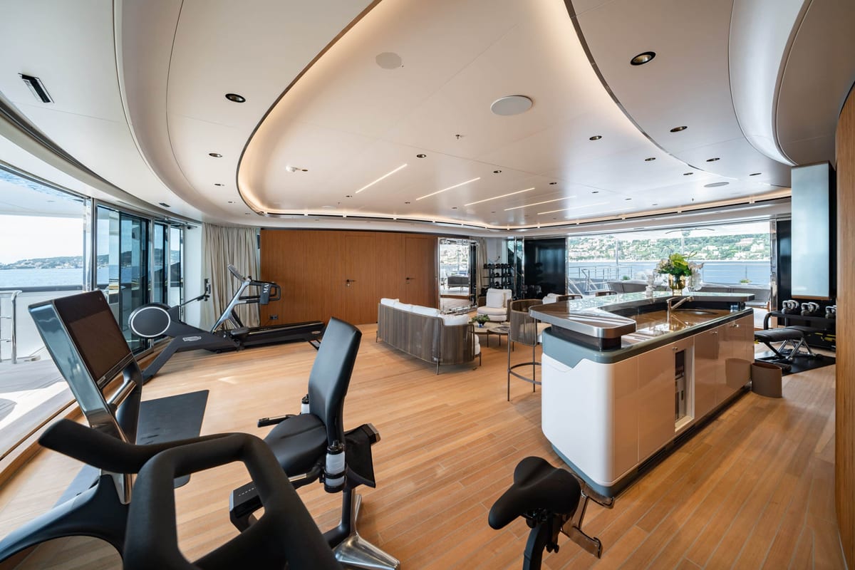 Superyacht interior gym with exercise bikes and treadmill beside a bar and lounge area.