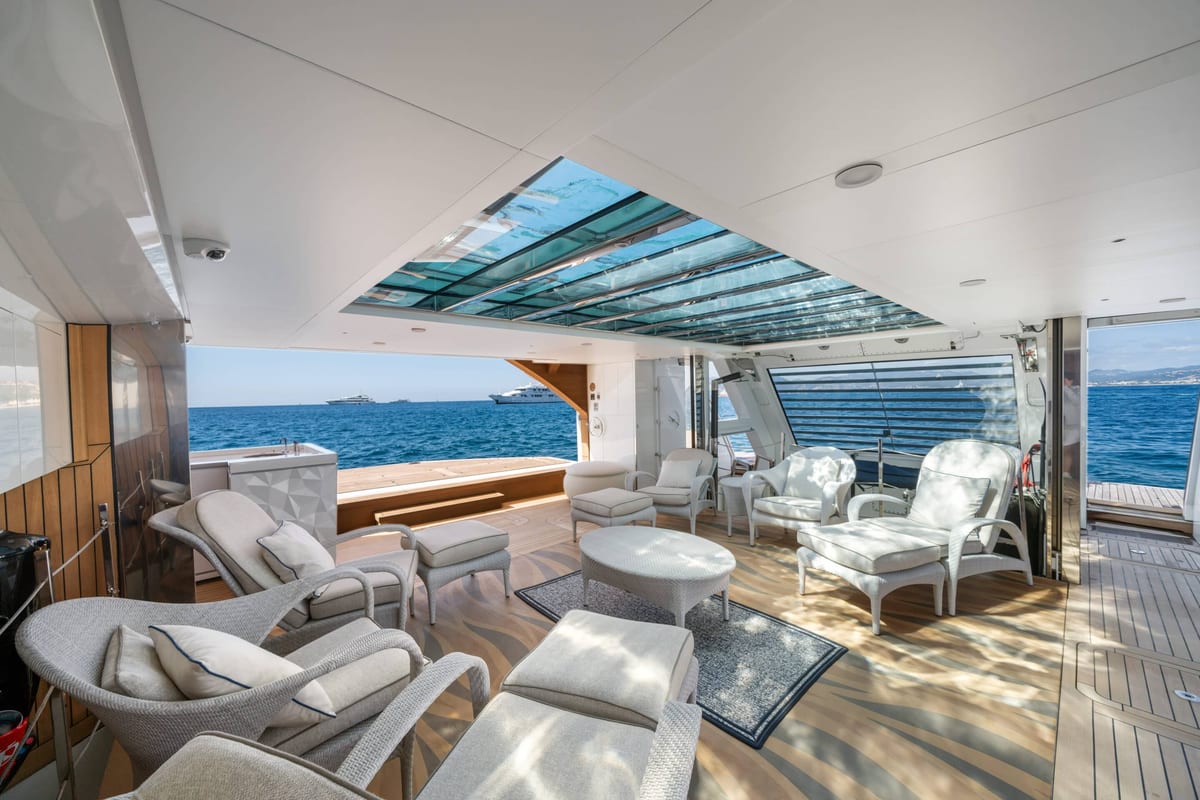 Shaded aft deck lounge on a yacht with wicker armchairs, glass skylight and open sea views.